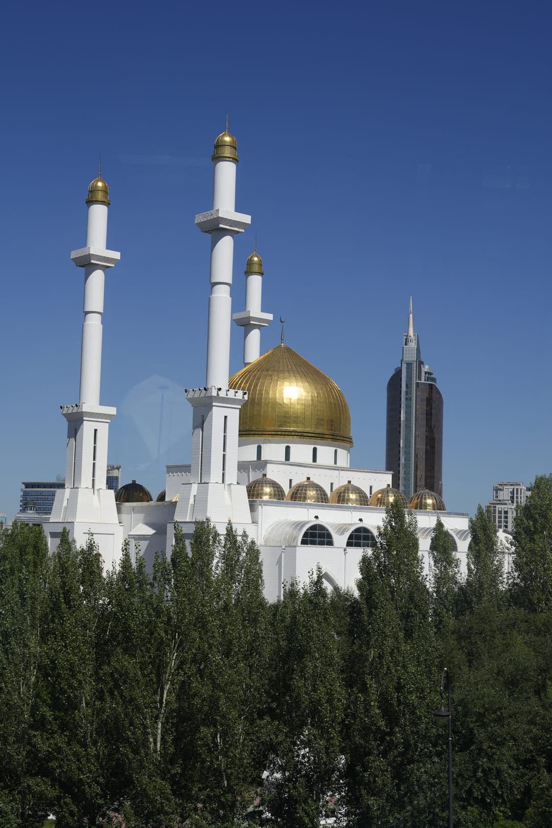 A white mosque with a golden central dome and four slim white minarets, surrounded by trees
