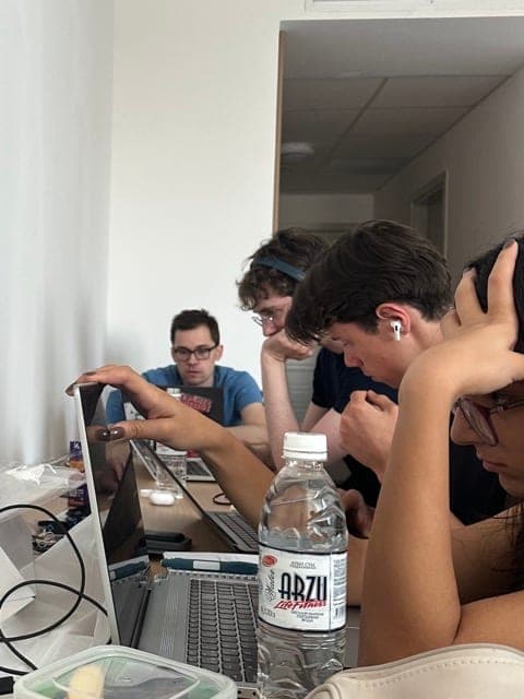 Four team members hunched over open laptops in a dorm room, focused on code