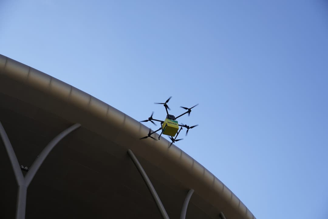 A hexacopter delivery drone with a yellow package flying past the curved roof of a building against a blue sky