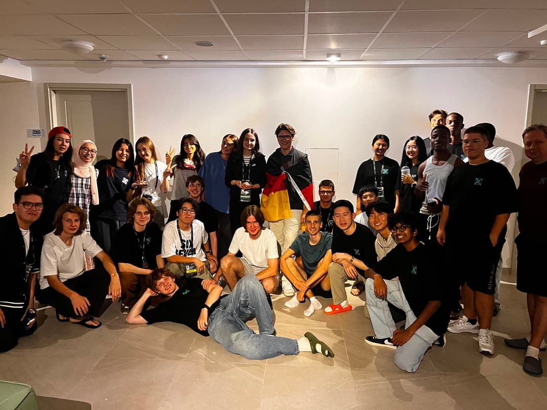 Large group photo of around twenty-five contestants from many different countries posing together in a common room, with a German flag in the middle