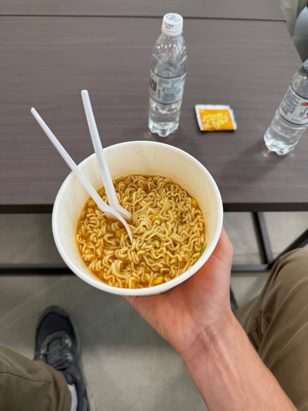 A paper cup of instant ramen held over a table, with two plastic forks sticking out