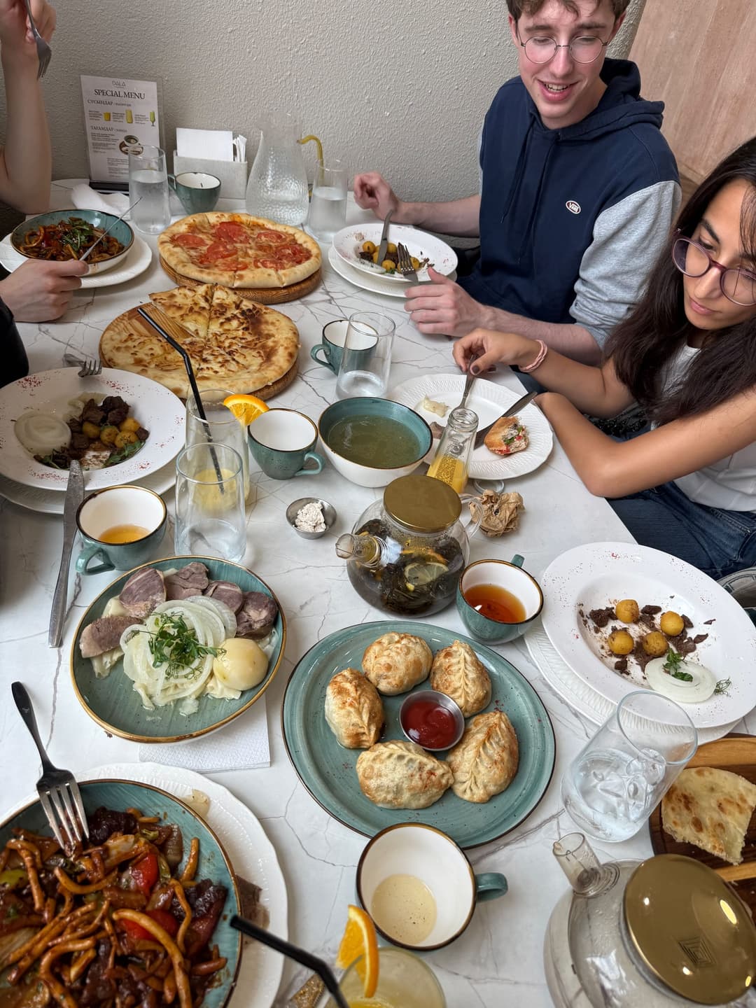 Table crowded with Kazakh dishes: horse meat, dumplings, soup and a margherita pizza