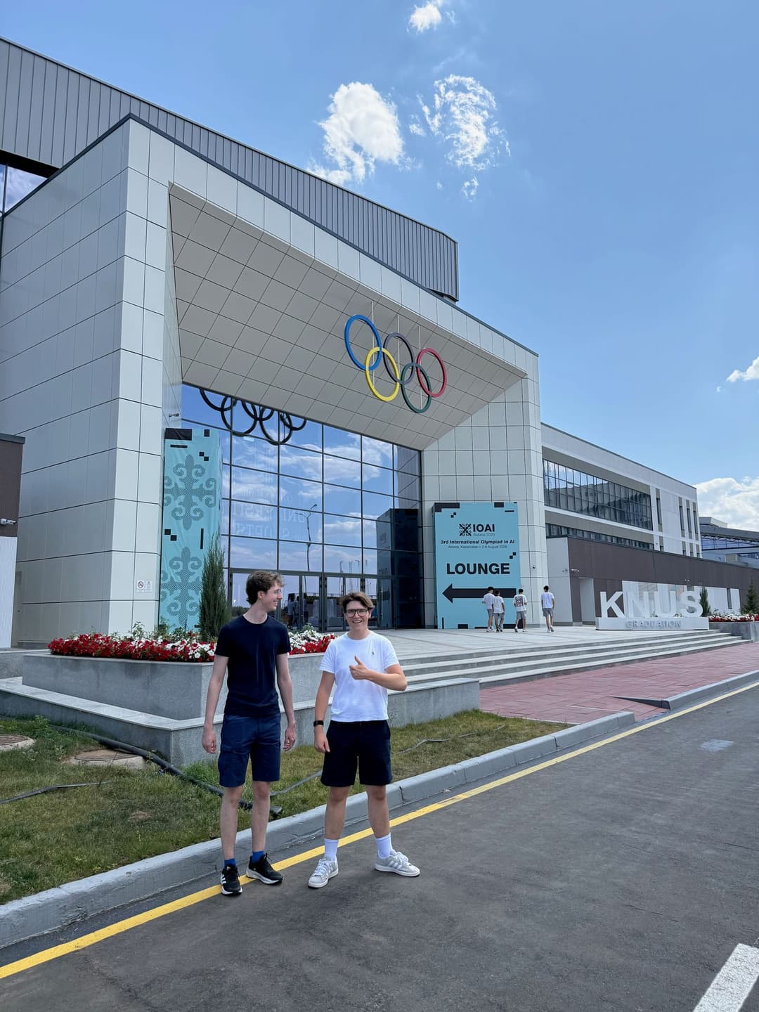 Two team members standing in front of the Kazakh National University of Sports, with the Olympic rings mounted on the building's glass facade