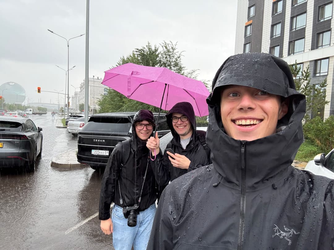 Selfie of three team members in rain jackets under heavy rain, one holding a pink umbrella