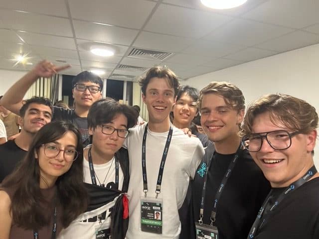 Group selfie of seven contestants from different countries pressed together in a dorm hallway, smiling on the last evening
