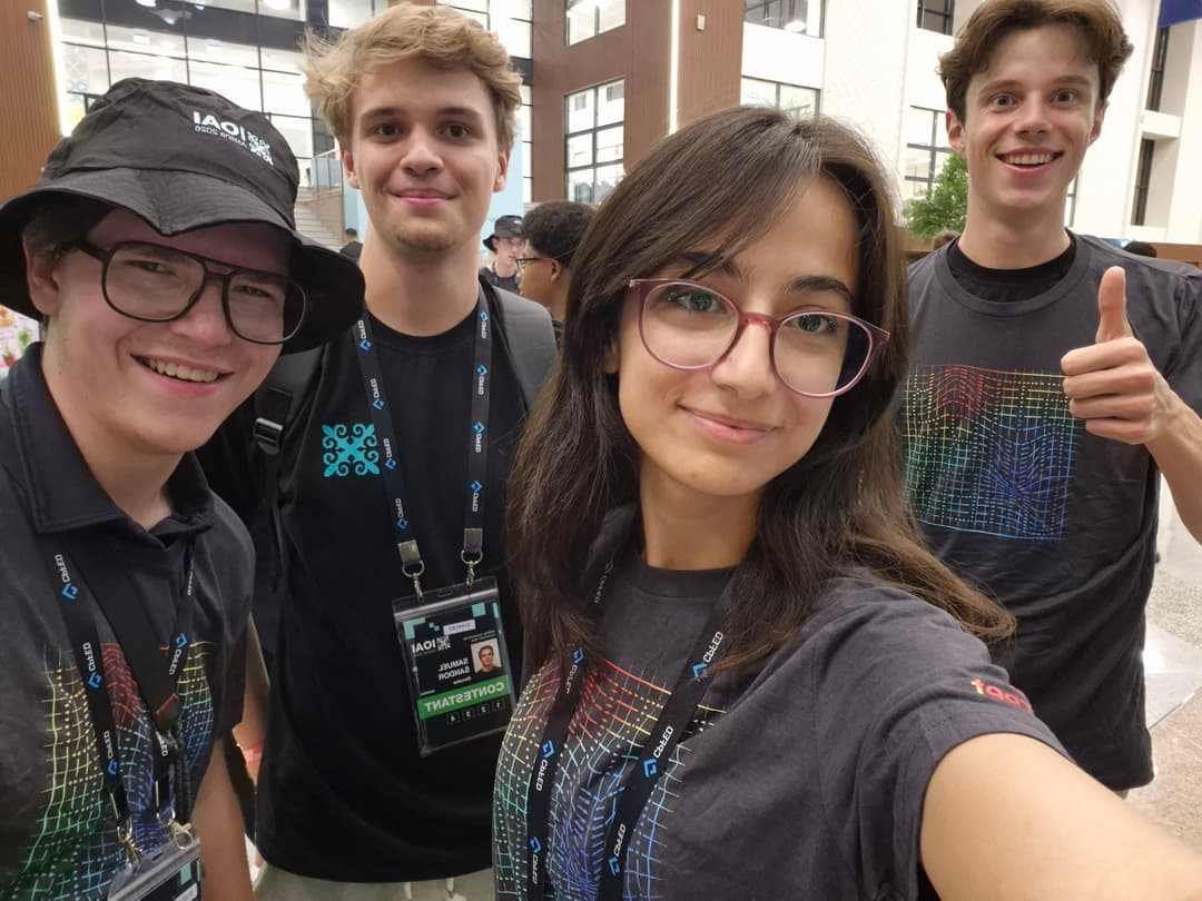 Selfie of four IOAI contestants in matching contest shirts and lanyards in the venue lobby
