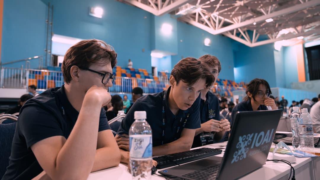 Three team members leaning in around a laptop showing the IOAI 2026 logo, deep in concentration during the team challenge